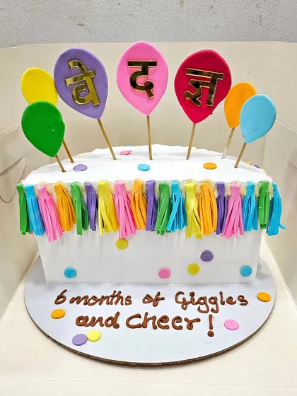 A colorful cake for a 6-month half-birthday celebration, decorated with bright tassels and balloon toppers.