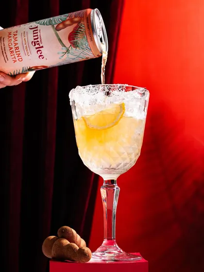 A dramatic pour shot for Junglee's Tamarind Margarita. The dark, theatrical background and the action of pouring into a salt-rimmed glass create a sophisticated and enticing image.
