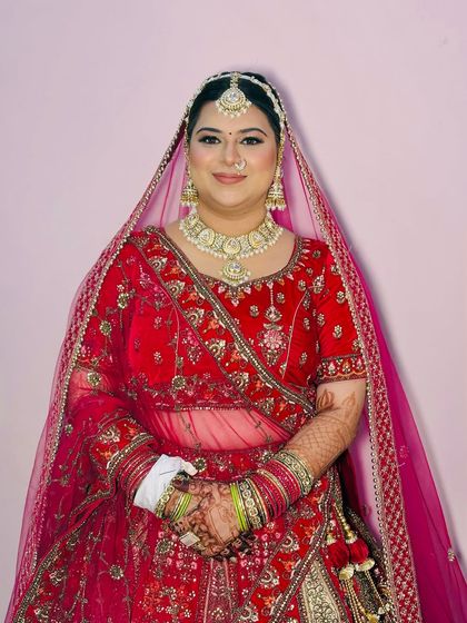 A full-length portrait of the bride, seated and smiling. The makeup is perfectly balanced with her heavy jewellery and ornate red lehenga.