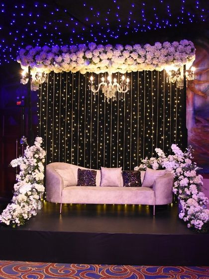 A cozy and glamorous seating area for a Sangeet. A plush couch is set against a backdrop of twinkling fairy lights, with a canopy of white flowers and a chandelier adding to the starry night ambiance.