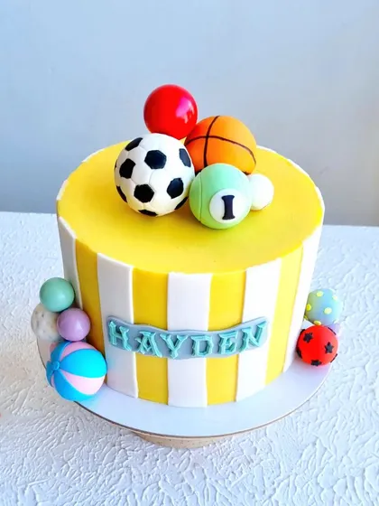 A colorful, ball-themed cake with yellow and white stripes. The toppers include a soccer ball, basketball, and pool ball, all sculpted from fondant.