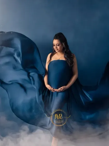 A celestial-themed maternity portrait. The mother-to-be is wrapped in flowing blue fabric against a night sky backdrop with a full moon, creating a serene and magical image.