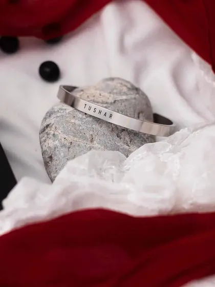 A silver cuff engraved with 'TUSHAR', placed on a stone with elegant white and red fabrics, highlighting its gift-worthy quality.