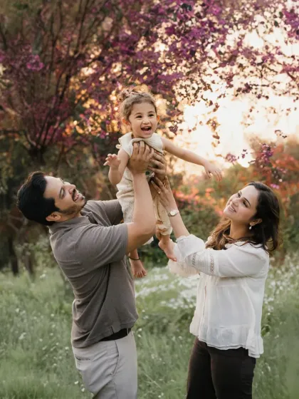 Parents lift their laughing daughter into the air against a backdrop of beautiful pink-flowered trees. This is a moment of pure, uninhibited family fun.