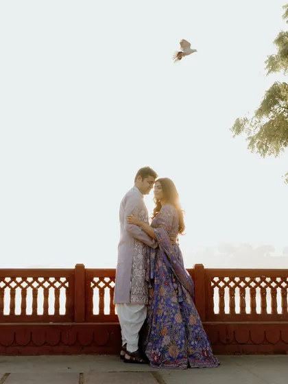 A couple embraces against a red stone railing as the sun sets behind them. A bird flies overhead, adding a touch of beautiful chance to this perfectly timed, romantic pre-wedding photo in Jaipur.