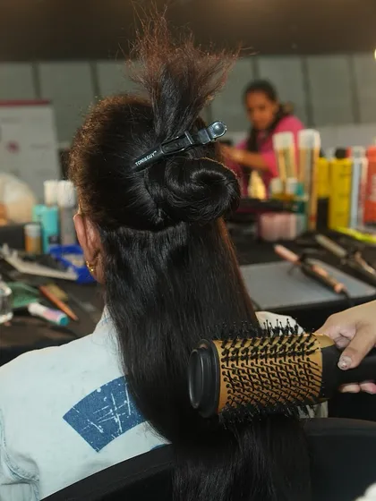 A student uses a round brush and blow dryer to style a model's long hair. Mastering blowouts is a fundamental skill for any hair artist.
