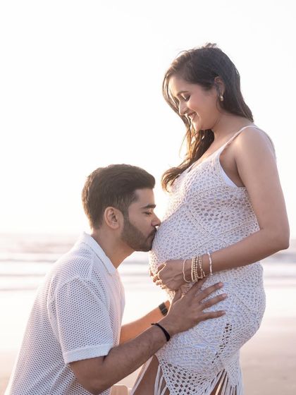 A classic maternity pose on the beach. The partner kneels to kiss the baby bump, with the ocean and golden hour light creating a beautiful, serene backdrop.