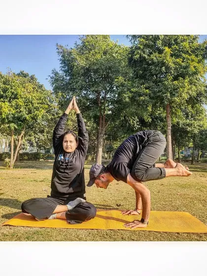 An instructor demonstrates Bakasana (Crow Pose) while a student holds Padmasana (Lotus Pose) during a sunny park session.
