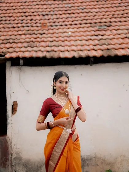 A smiling portrait of a model in a traditional saree against a rustic village house. The composition creates a warm and inviting atmosphere.