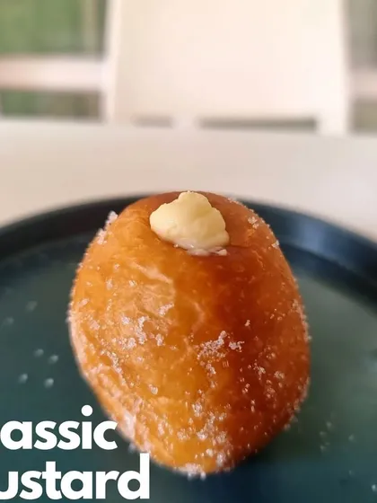 A close-up of our Classic Custard Bomboloni. The soft, sugar-dusted doughnut is filled with a smooth and luscious vanilla custard, a timeless favorite.