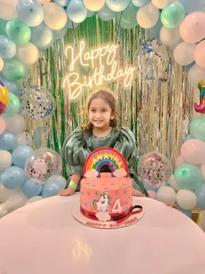The birthday girl smiling proudly with her unicorn and rainbow-themed 4th birthday cake. The pastel balloon arch completes the magical scene.