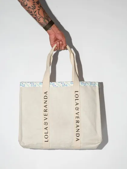 A tattooed arm holds up a canvas tote bag against a stark white background with dramatic lighting. This is a clean, edgy shot that clearly displays the product for an online store.