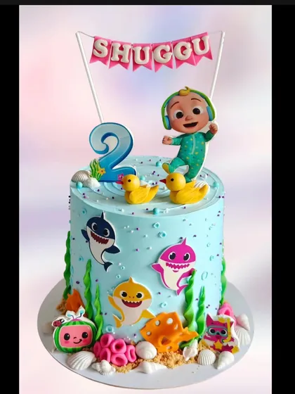 A Cocomelon and Baby Shark combination cake. This design brings together two of the most popular themes for toddlers, featuring JJ and the friendly sharks in a colorful underwater setting.