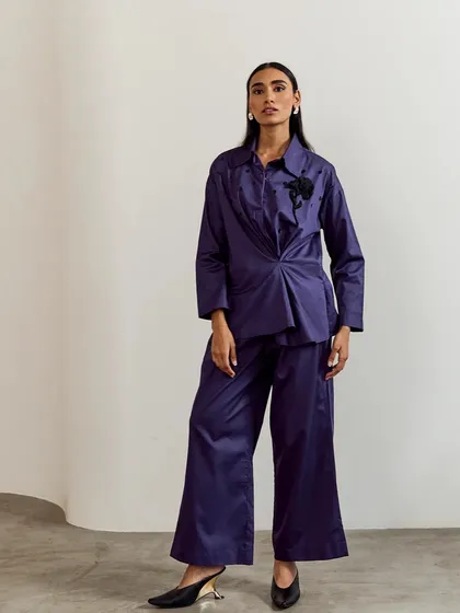 For the modern woman, this deep purple co-ord set offers a powerful statement. The structured top with its unique twist detail and wide-leg trousers create a silhouette that is both minimalist and maximalist.