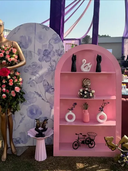 A creative and artistic installation for a Haldi, featuring a mannequin adorned with a dress made of fresh pink roses and a pink shelving unit with curated decor items.