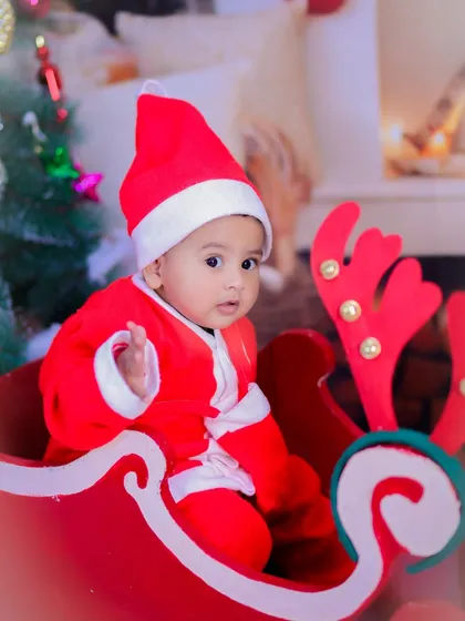 A close-up of a baby in a Santa suit, looking curiously at the camera from a festive red sleigh. A perfect holiday portrait.