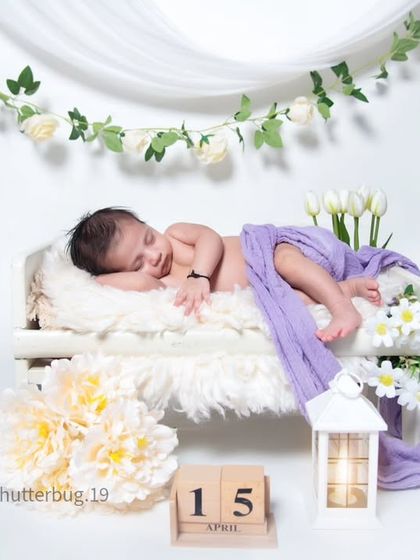 A simple and elegant setup with a clean white background. The baby sleeps on a miniature bed, draped in a lavender cloth, with white flowers and a garland adding a touch of nature.