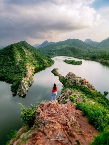 A stunning drone photograph of Bahubali Hills in Udaipur. This aerial view captures a person standing on the cliff's edge, providing a sense of scale against the vast, beautiful lake.