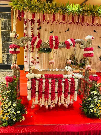 A classic naming ceremony setup with a decorated cradle on a red carpet. The backdrop of hanging jasmine strings and a floral canopy creates a traditional and festive atmosphere.