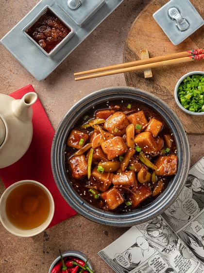 A tofu dish in a dark sauce, styled with manga-themed paper for YouMee, reflecting the restaurant's quirky and modern brand identity.