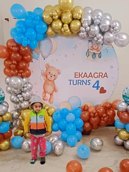 A sweet teddy bear theme for a 4th birthday. The backdrop features a cute bear illustration, surrounded by a balloon arch in brown, blue, gold, and silver.