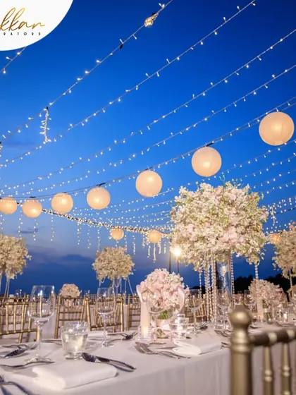 A beautiful al fresco dining setup for a reception. The tables are set under a canopy of string lights and paper lanterns, creating a magical and romantic atmosphere against the evening sky.