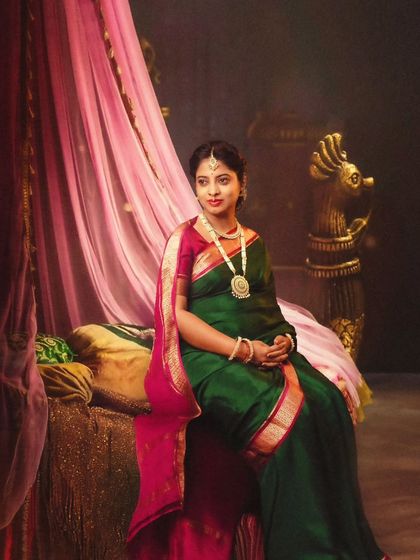 A regal portrait of a mom-to-be in a rich green and pink saree, seated on a throne-like chair in a palatial setting.