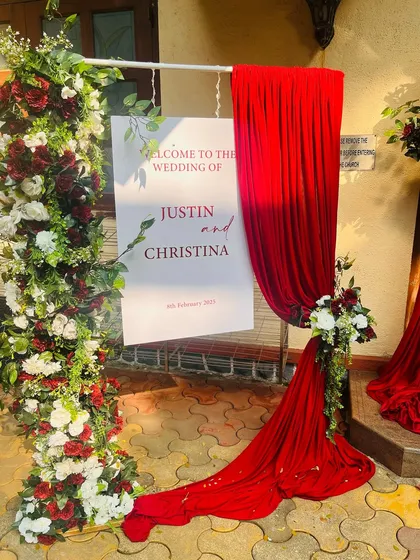 A personalized welcome sign for a wedding, elegantly styled with a floral pillar and flowing red fabric. This is the first detail guests see, setting the tone for the event.