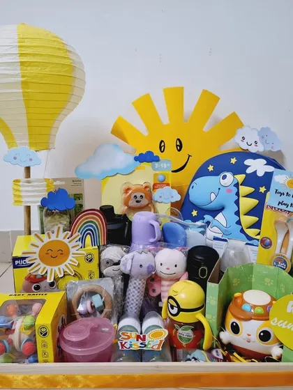 This 'Hello Sunshine' hamper is all about fun and play. It's filled with a variety of toys, including rattles, a Minion figure, a dinosaur pillow, and a yellow hot air balloon lantern.