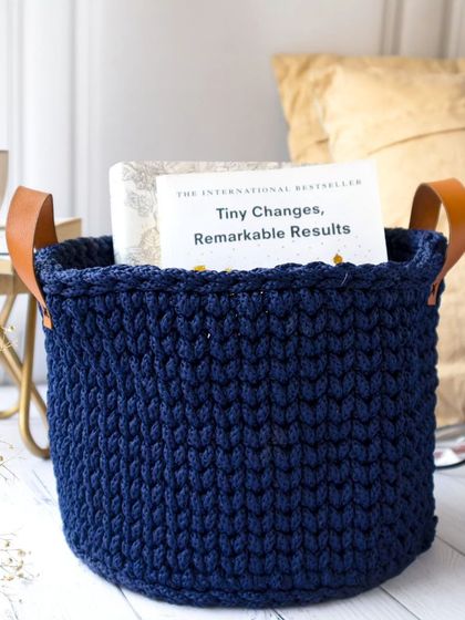 A cozy reading nook scene featuring a hand-crocheted navy blue storage basket holding a book. The soft lighting and comfortable setting highlight the basket's practical and aesthetic use in home organization.