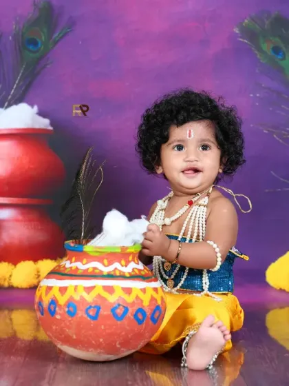 Another angle of the curly-haired Little Krishna, sitting happily among the festive props. This shot captures the joyful and mischievous spirit of the theme.