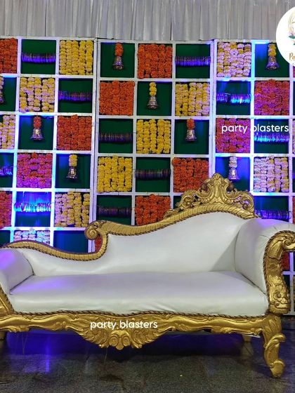 A traditional baby shower backdrop, featuring a grid of fresh marigold flowers and temple bells. The ornate gold and white chaise lounge completes the regal and festive look.