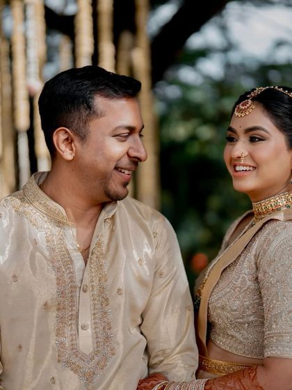 A close-up of a smiling couple. His cream silk kurta with fine embroidery on the neckline complements her fully embroidered gold blouse and saree, showcasing our attention to detail in couple's wear.