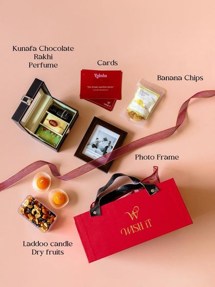 A flat lay of the contents of our red Rakhi hamper. It includes a watch box, Kunafa chocolate, perfume, a resin Rakhi, a photo frame, and more.