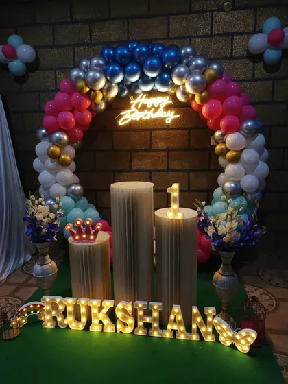 This first birthday setup against a brick wall features a circular balloon arch in pink, blue, and silver. A neon sign and light-up marquee letters add a warm glow to the celebration.