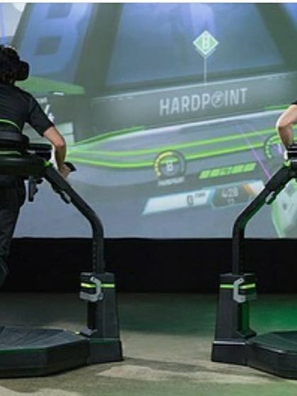 Another angle of the two-player Virtuix Omni setup, with the game projected on a large screen for spectators to follow the action.