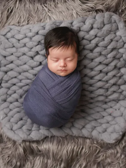 A newborn swaddled in a deep blue wrap, resting on a grey knit blanket and fur. The cool tones create a serene and calming mood for this infant portrait.