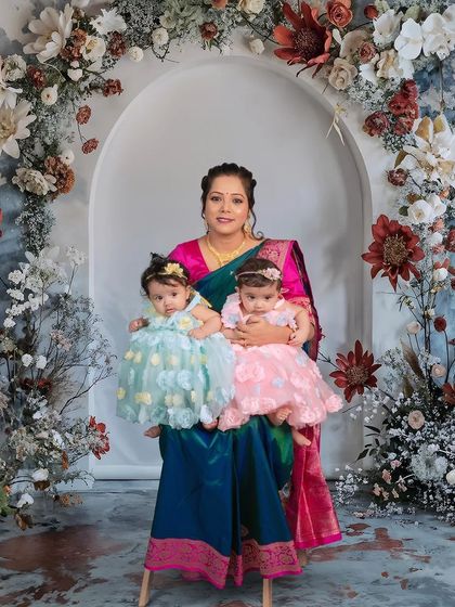 A proud mother holds her twin daughters in this beautiful portrait. The intricate floral backdrop complements her traditional saree, making for a truly artistic and heartfelt image.
