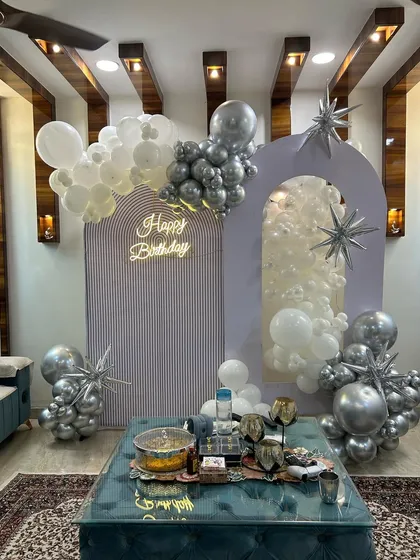 A modern and chic birthday setup in a living room. This design features elegant arched backdrops, a neon sign, and balloon clusters in a sophisticated silver and white color scheme.