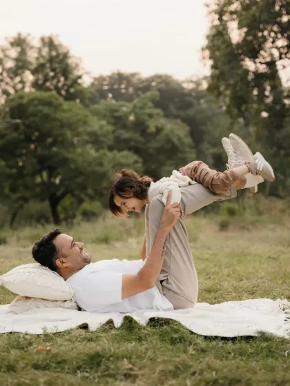 A father lies on the grass and playfully lifts his child with his feet. This fun "airplane" pose is a classic for father and child photoshoots.