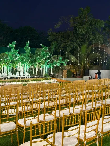 A wide view of the enchanted forest reception setup. The golden guest chairs lead the eye towards the magical stage, with fairy lights twinkling in the trees above, creating a cohesive and immersive environment.