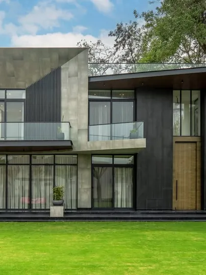 This farmhouse facade uses an earthy, grey-toned sustainable design to blend with its lush surroundings. The structure combines bold concrete forms with dark metal cladding and large glass windows for a perfect harmony between modern elegance and eco-conscious living.