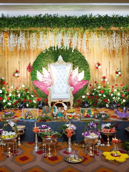 A clear, full view of a traditional stage decoration. The design is centered around a white Maharaja chair against a lotus-themed backdrop, framed by greenery and floral arrangements, with all the ceremonial trays neatly presented.