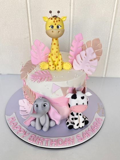 A soft and sweet jungle animal cake with a pink and pastel theme, featuring a giraffe, elephant, and cow surrounded by tropical leaves.