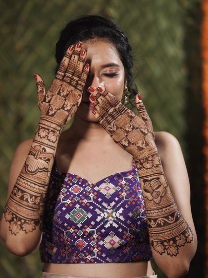 A happy bride posing with her full-sleeve mehendi. The intricate design looks stunning with her purple and white outfit.