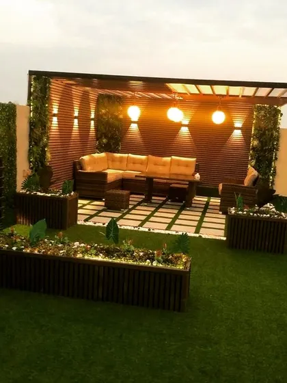 This pergola creates a complete outdoor room with an L-shaped sofa and planters framing the space. The back wall features wood paneling and is lit by multiple sconces, creating a warm and inviting focal point.