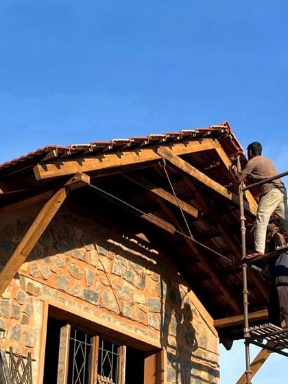 Workers carefully place Mangalore tiles on the roof of a stone house. The warm sunlight highlights the texture of the stone and the richness of the wood, even during construction.