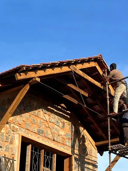 Workers carefully place Mangalore tiles on the roof of a stone house. The warm sunlight highlights the texture of the stone and the richness of the wood, even during construction.