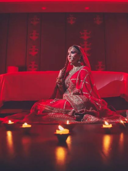 A bride in a red lehenga sits on the floor, surrounded by diyas, creating a warm and traditional portrait.