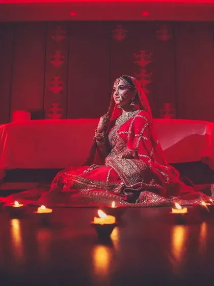 A bride in a red lehenga sits on the floor, surrounded by diyas, creating a warm and traditional portrait.
